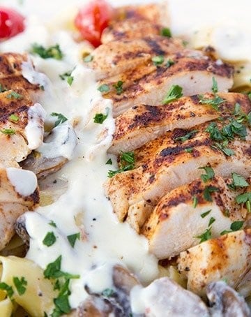 slices of seasoned grilled boneless skinless chicken breast sitting on top of Pappardelle with alfredo sauce, grilled cherry tomatoes and mushrooms sprinkled with chopped parsley