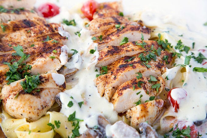 close up of slices of seasoned grilled boneless skinless chicken breast sitting on top of Pappardelle with alfredo sauce, grilled cherry tomatoes and mushrooms sprinkled with chopped parsley