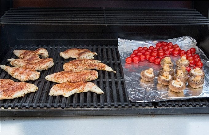 seasoned chicken breasts on a barbecue grill, with cherry tomatoes and mushrooms on foil next to the chicken