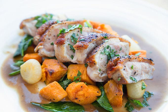 slices of cooked duck breast on a bed of sweet potato chunks, spinach leaves and pearl onions on a white plate with a brown gravy around the edges of the plate