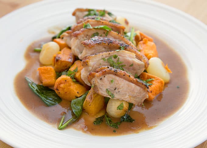 slices of pan seared duck breast on a bed of sweet potato chunks, spinach leaves and pearl onions on a white plate with a brown gravy around the edges of the plate