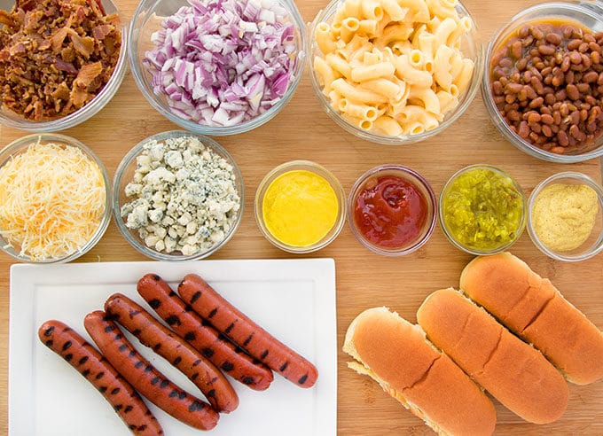 bowls of toppings for a hot dog bar with grilled ball park hot dogs on a white platter with hot dog rolls next to it all sitting on a wooden cutting board