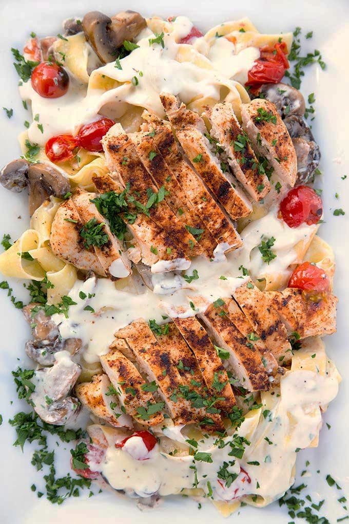 2 grilled chicken breasts cut into slices sitting on top of Pappardelle with alfredo sauce, grilled cherry tomatoes and mushrooms sprinkled with chopped parsley on a white serving platter