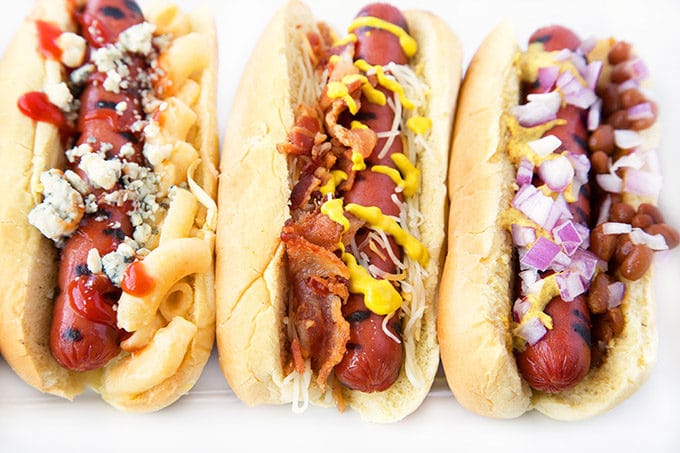 Grilled Hot Dogs on Potato Rolls with different toppings sitting on a White Platter