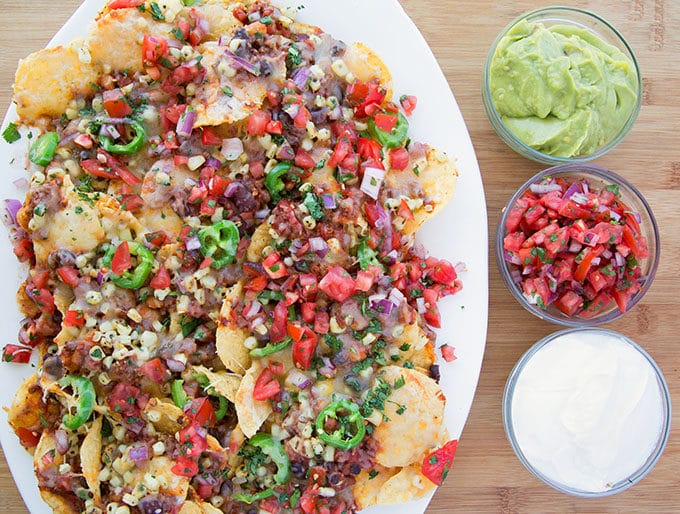 ultimate nachos loaded with fresh salsa, jalepeno slices, chili and cheese on a white oval platter. Sitting on a wooden cutting board with bowls of guacamole, sour cream and salsa fresca