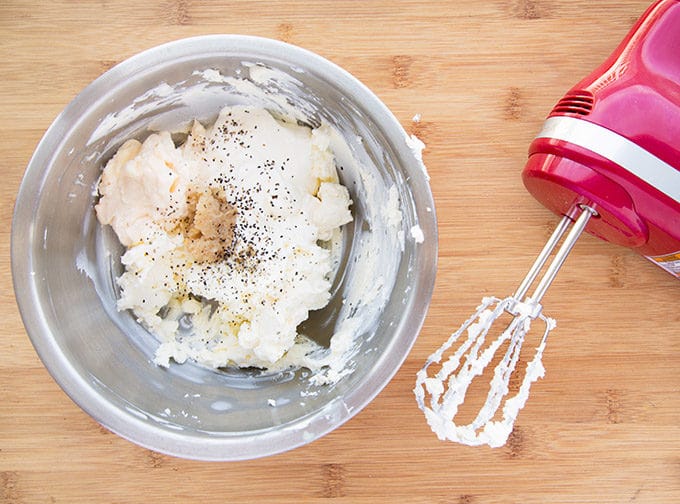 cream cheese in stainless steel bowl with other ingredients in the bowl, sitting on a wooden cutting board next to a red electric hand mixer