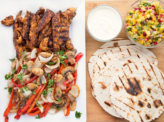a rectangular white platter with seasoned grilled chicken strips and grilled sliced red peppers, onions and mushrooms next to a small glass bowls of lime crema, pineapple-mango salsa, and grilled tortillas sitting on a wooden cutting board