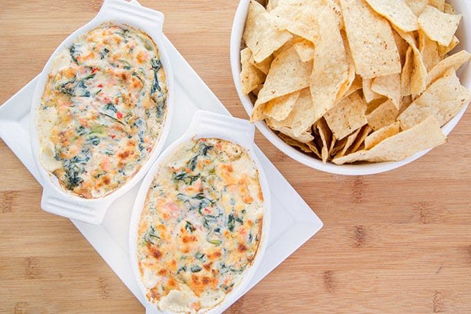 two white oval baking dish filled with salmon dip on a white tray next to a white bowl of tortilla chips all sitting on a wooden cutting board