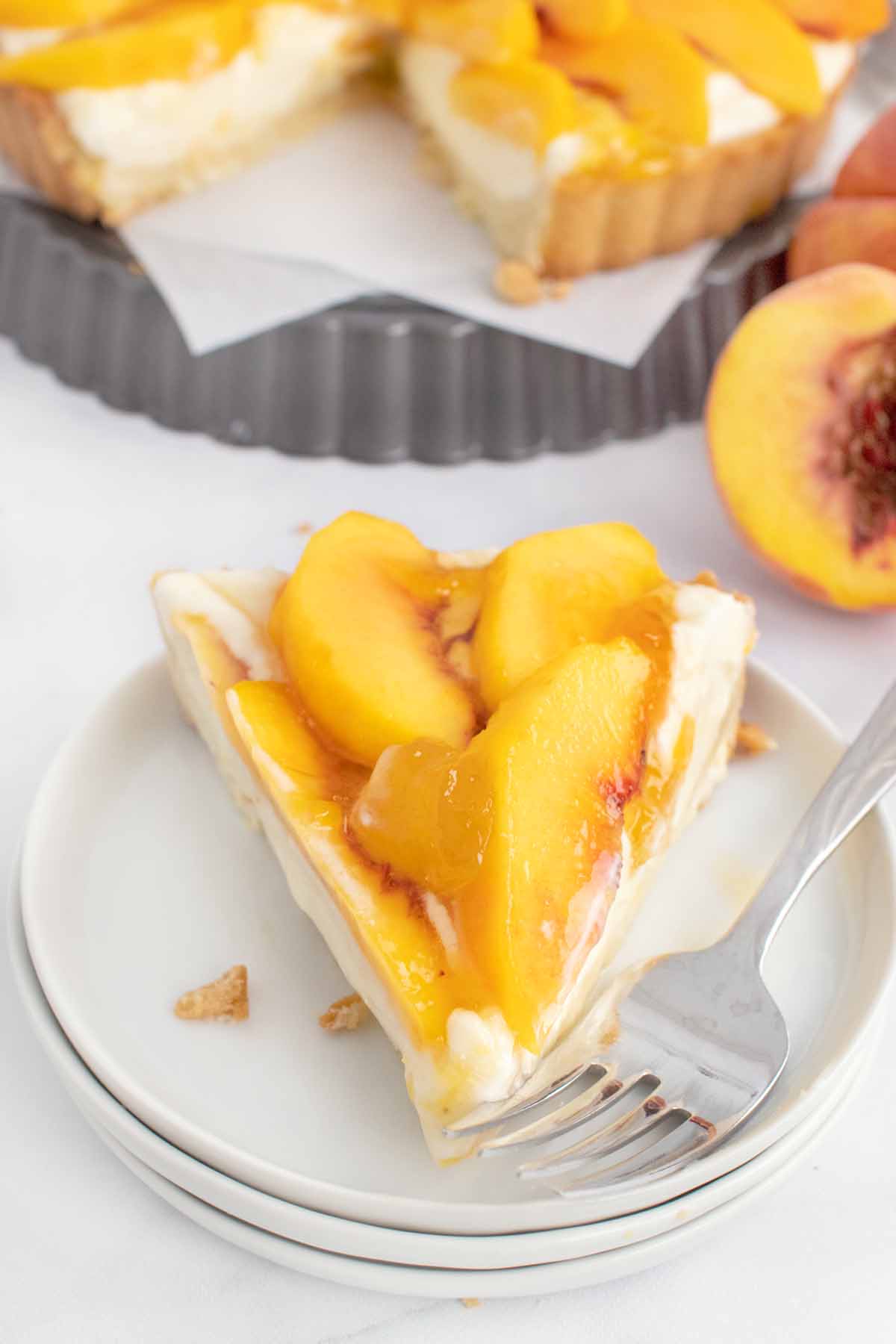 slice of peach tart on white plate with a fork with whole pie behind it