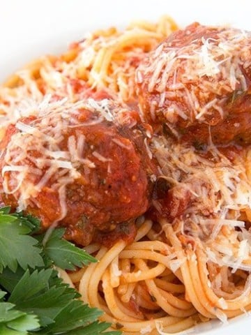 white bowl of spaghetti with two meatballs topped with grated romano cheese, with a sprig of Italian parsley in the bottom left corner