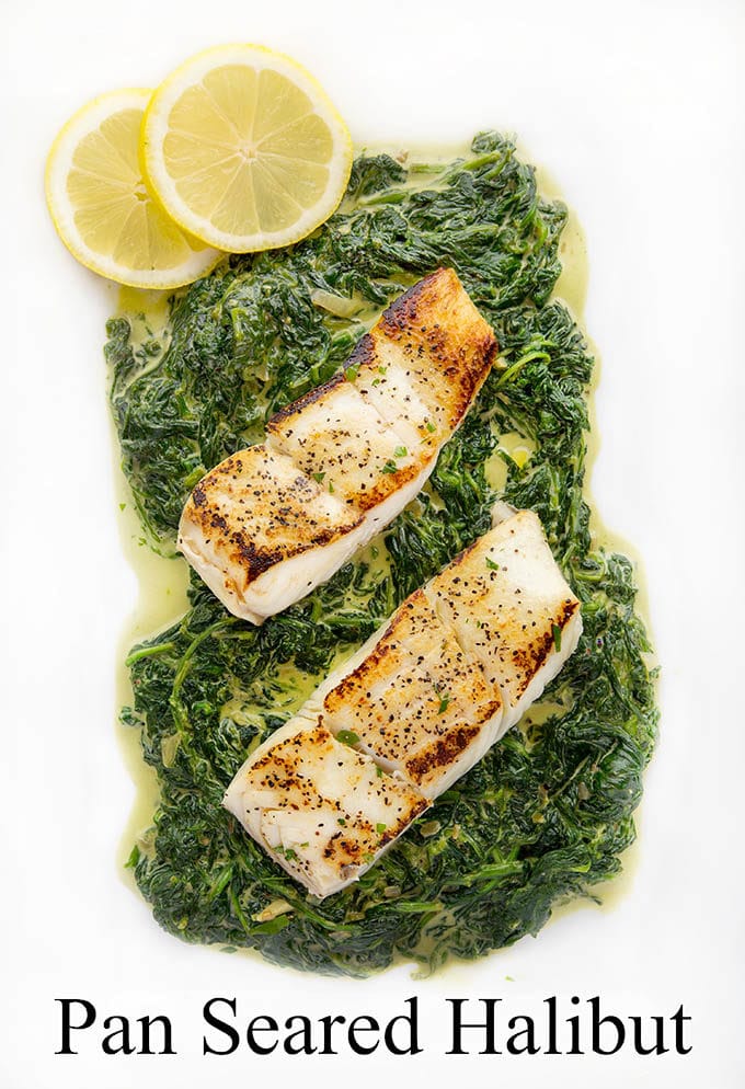 2 fillets of pan seared halibut sitting on a bed of creamed spinach on a white platter with 2 lemon slices in the upper left hand corner