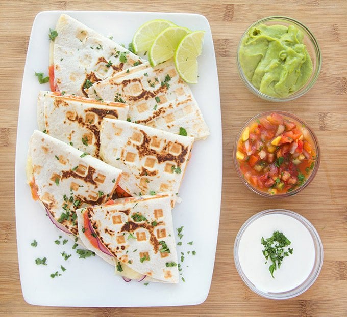 overhead view of smoked salmon quesadillas cut in half sitting on a white plate with a cilantro garnish sitting on a wooden cutting board with bowls of salsa, crema and guacamole