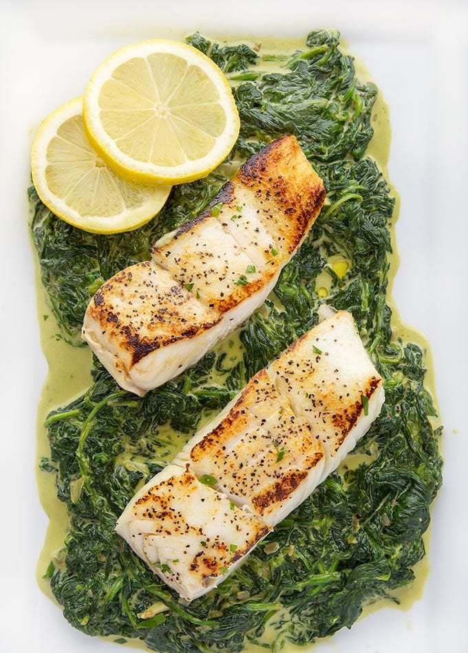 2 fillets of pan seared halibut sitting on a bed of creamed spinach on a white platter with 2 lemon slices in the upper left hand corner