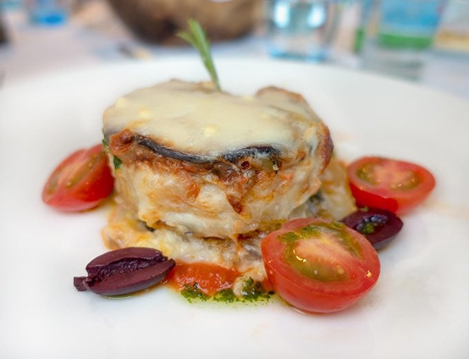 serving of eggplant moussaka on a white plate with cherry tomato halves around the eggplant