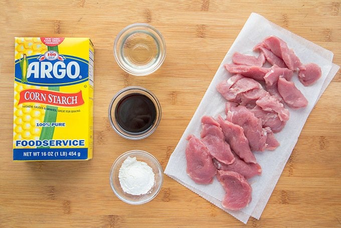 ingredients in small bowls next to sliced pork tenderloin on white butcher paper next to a box of Argo corn starch on a wooden cutting board