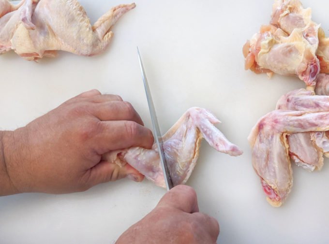one hand holding chicken wing while the other hand holds a chefs knife cutting thru the joint of the wing.