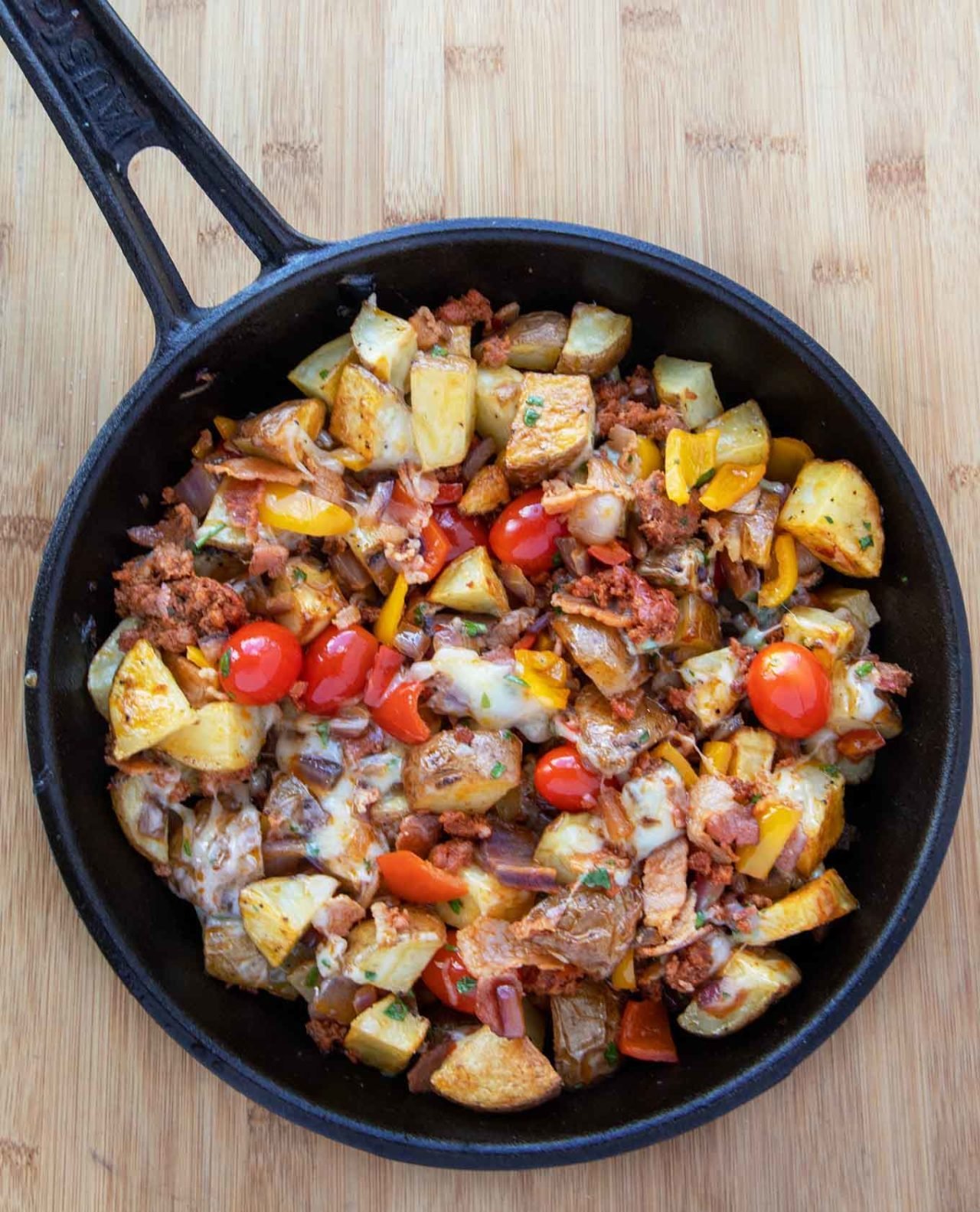 cast iron skillet filled with potatoes, sausage, bacon, onions, tomatoes, peppers and melted cheddar