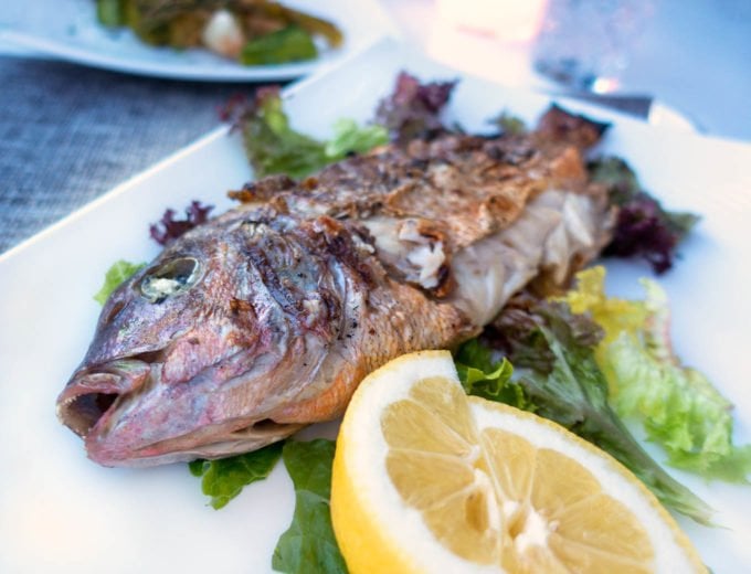 Whole cooked sea bass on a white plate garnished with lemon and greens