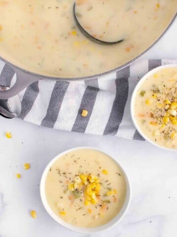 two white bowls of corn chowder with the pot of chowder and a grey and white striped towel