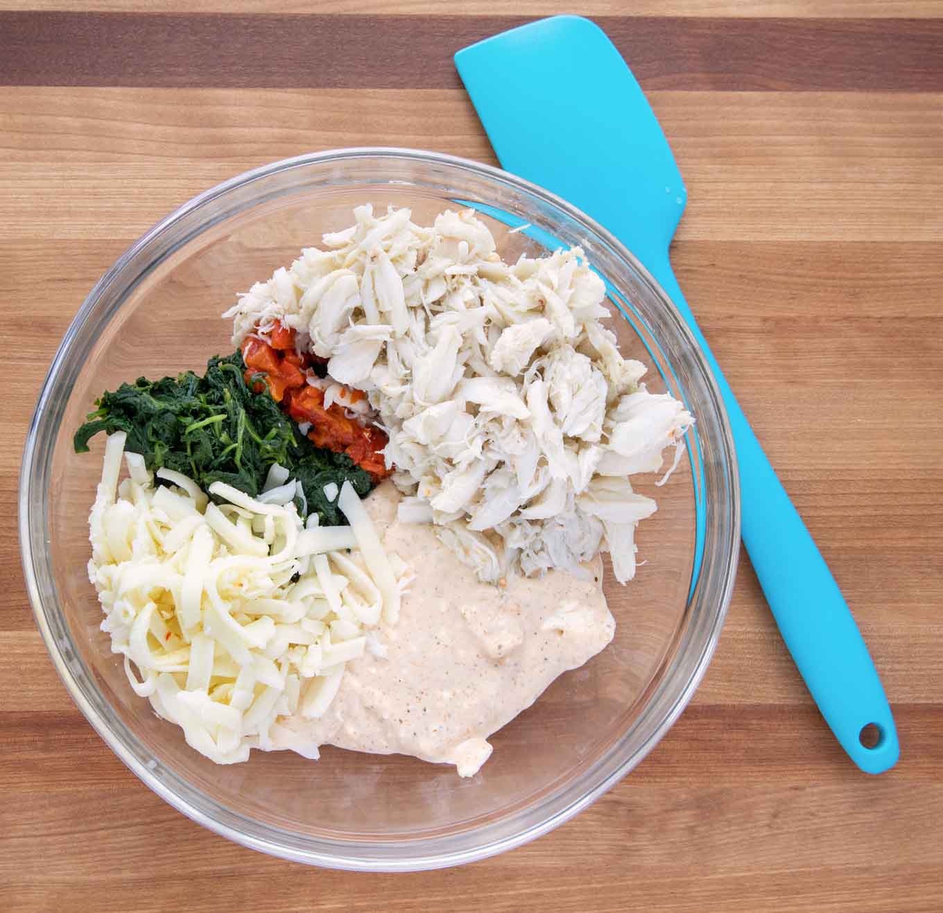 ingredients to make crabmeat and cheese stuffing in a glass bowl with a blue rubber spatula next to it