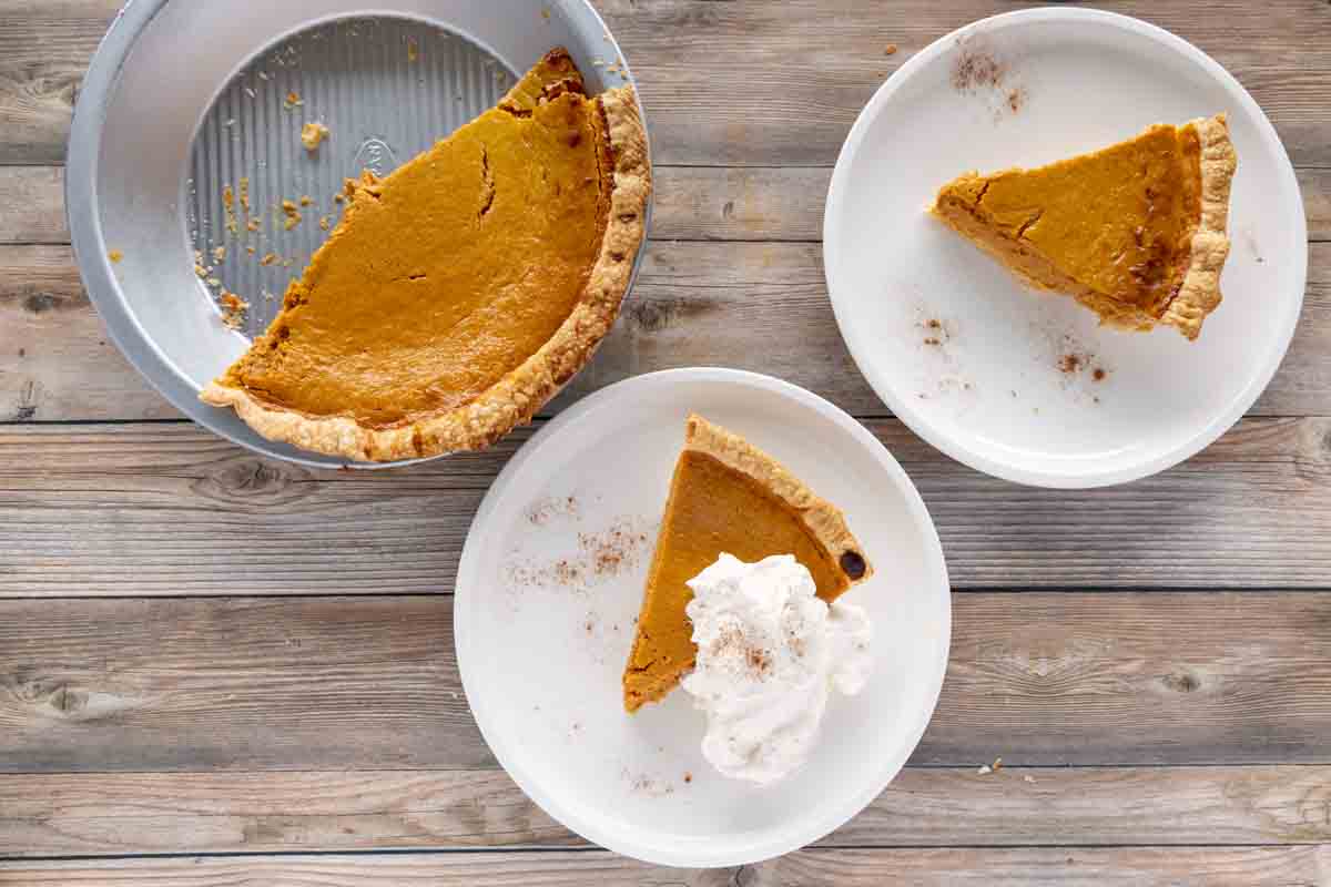 two slices of pumpkin pie on white plates with the rest of the pie in a metal pie pan