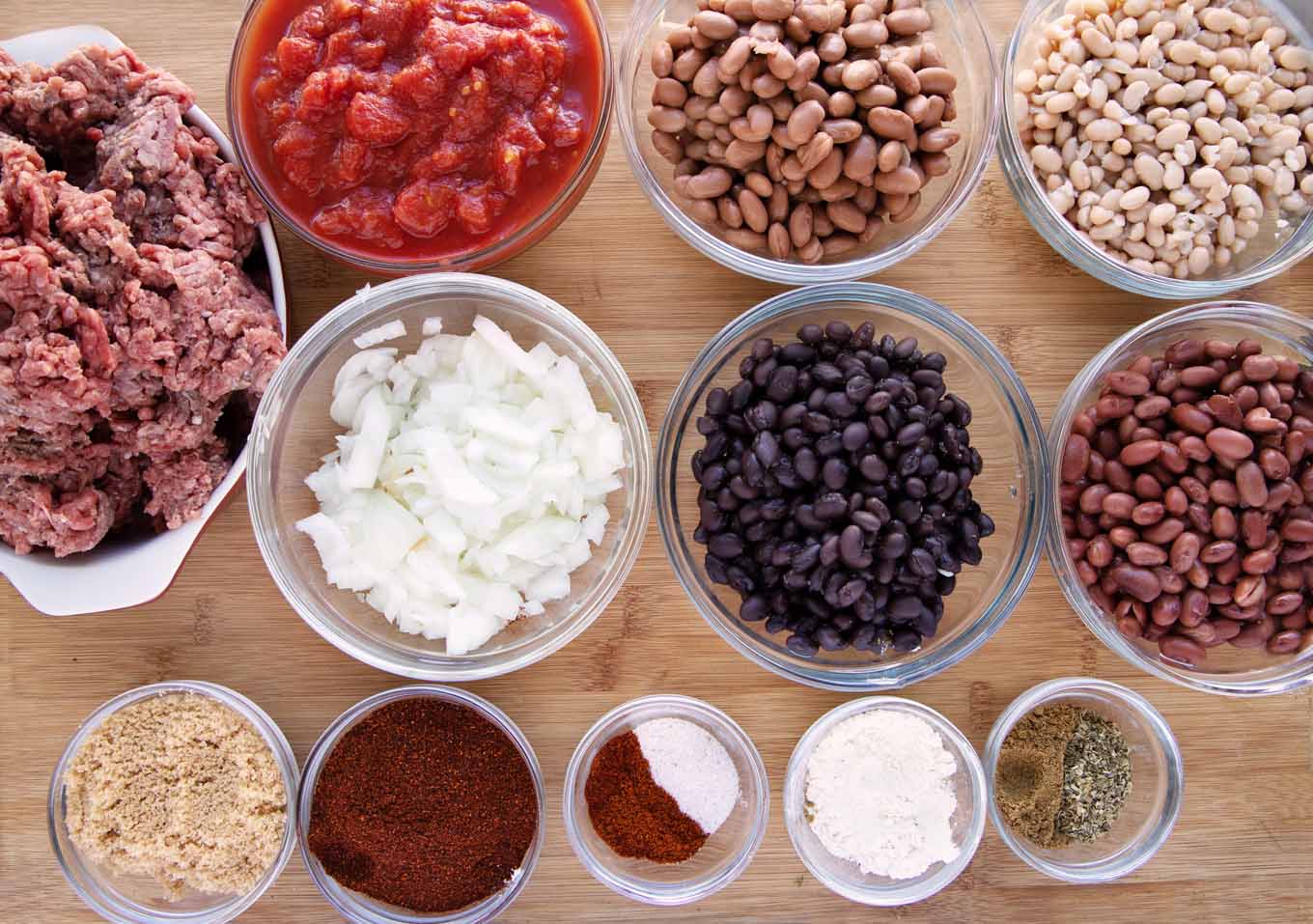ingredients to make four bean chili in glass bowls on a wooden cutting board