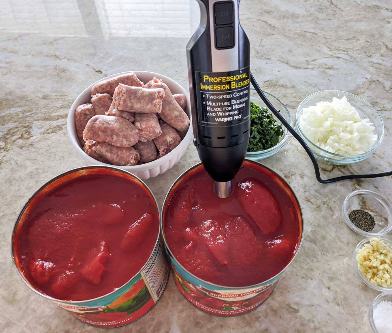 emersion blender in a #10 can of tomatoes with ingredients to make spaghetti sauce on a granite counter top