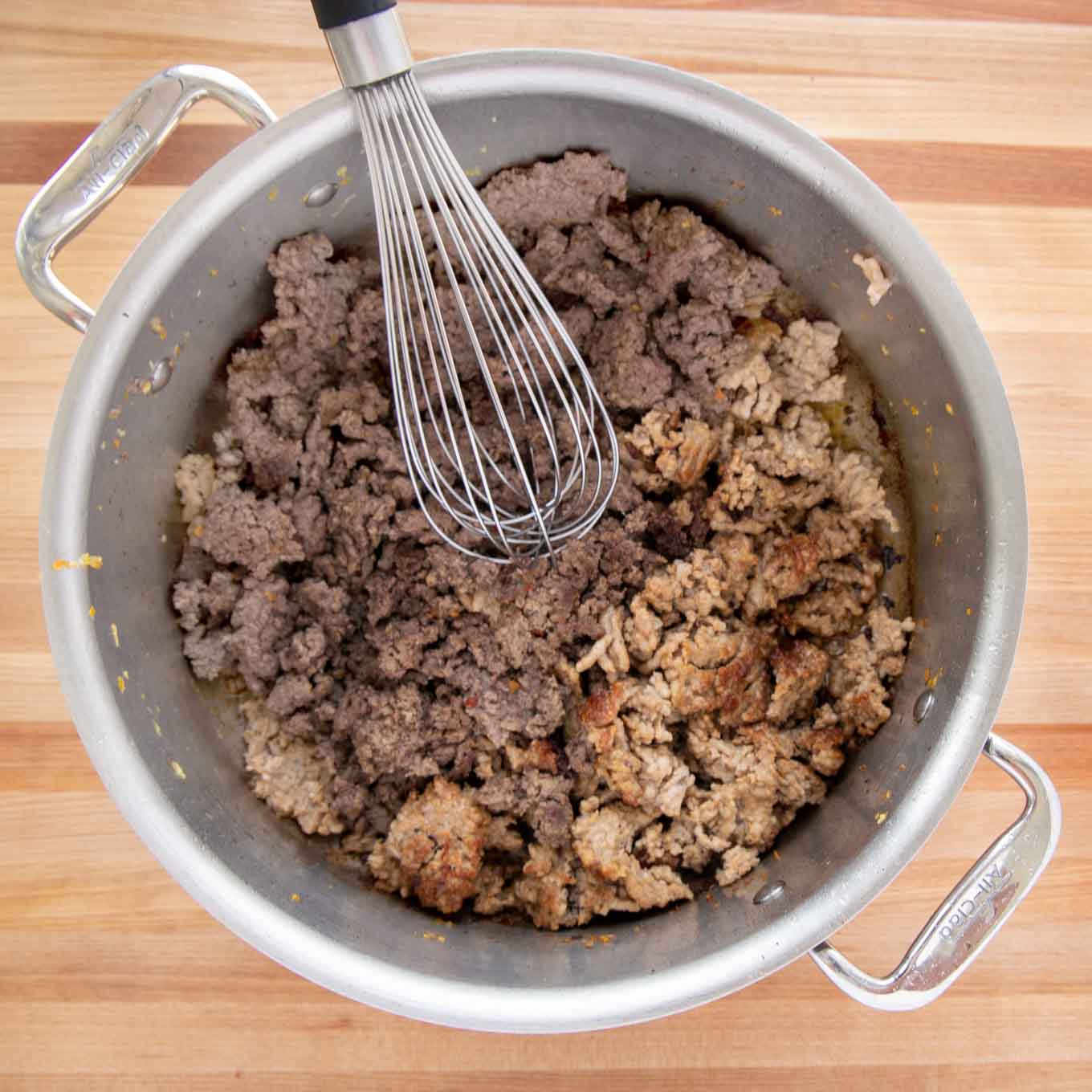 Beef and sausage cooked in a large pot with a wire whip in the pot.