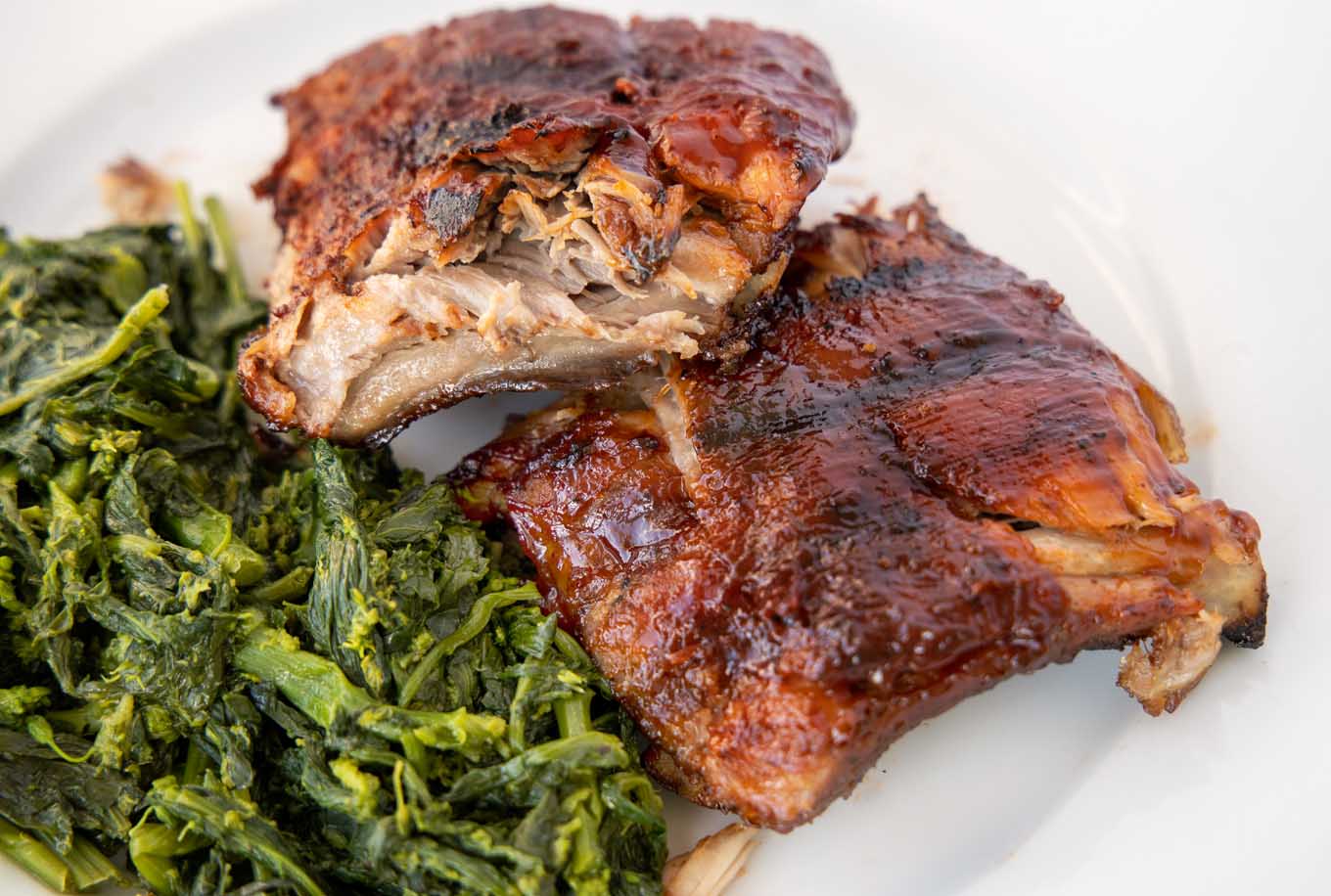 white plate with half rack of grilled baby back ribs cut in half and stacked, next to cooked broccoli rabe