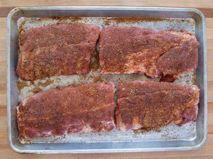 4 half racks of baby back ribs coated with a dry rub resting on a sheet pan