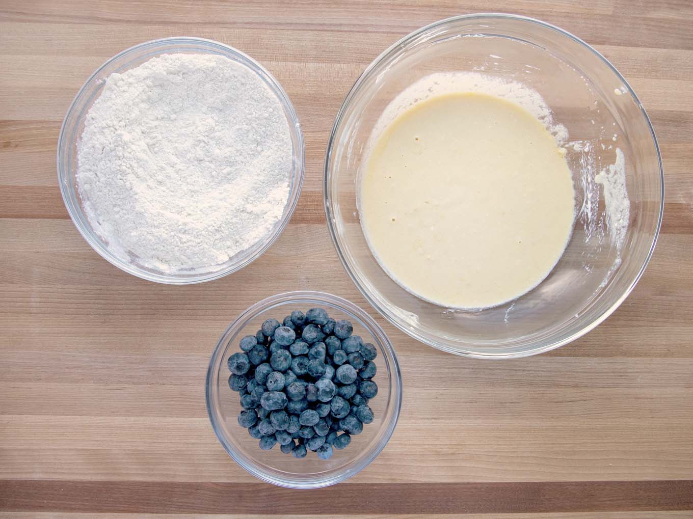 Glass mixing bowls with the flour mixture, egg mixture and blueberries on a wooden cutting board