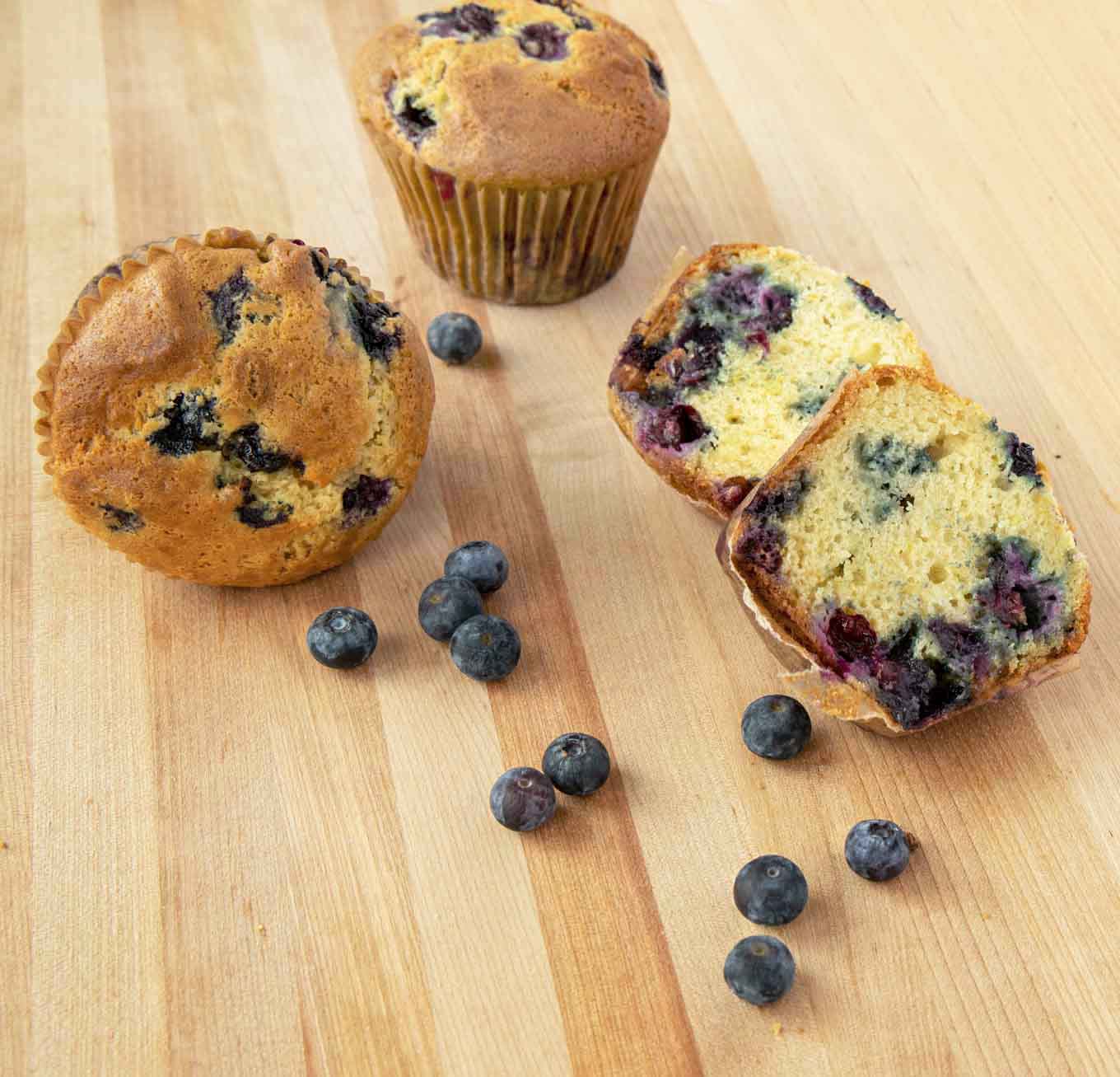 2 blueberry muffins and one muffin cut in half on a wooden cutting board with blueberries
