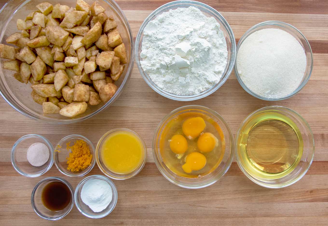 overhead view of the ingredients to make Jewish apple cake in glass bowls on a wooden cutting board