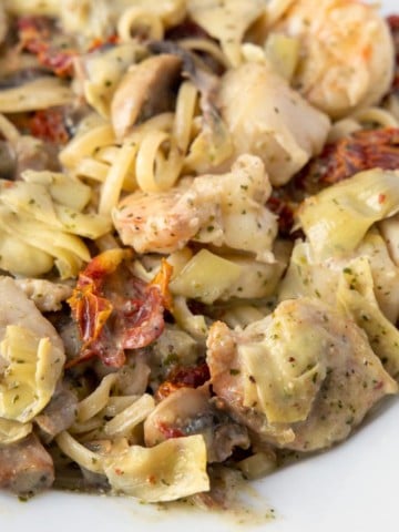 shrimp and scallops in a pesto sauce with sundried tomatoes, mushrooms and artichoke hearts over linguine in a white bowl