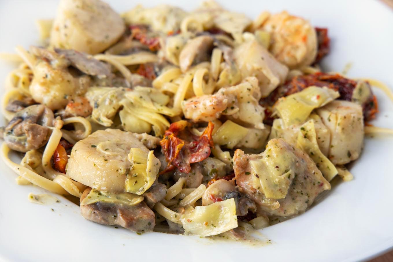 side view shot of seafood riviera consisting of shrimp and scallops in a pesto sauce with sundried tomatoes, mushrooms and artichoke hearts over linguine in a white bowl