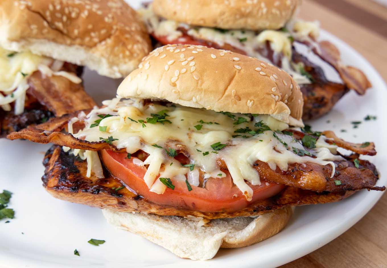 grilled chicken sandwich with bacon and tomato on a seeded bun with two more sandwiches in the background