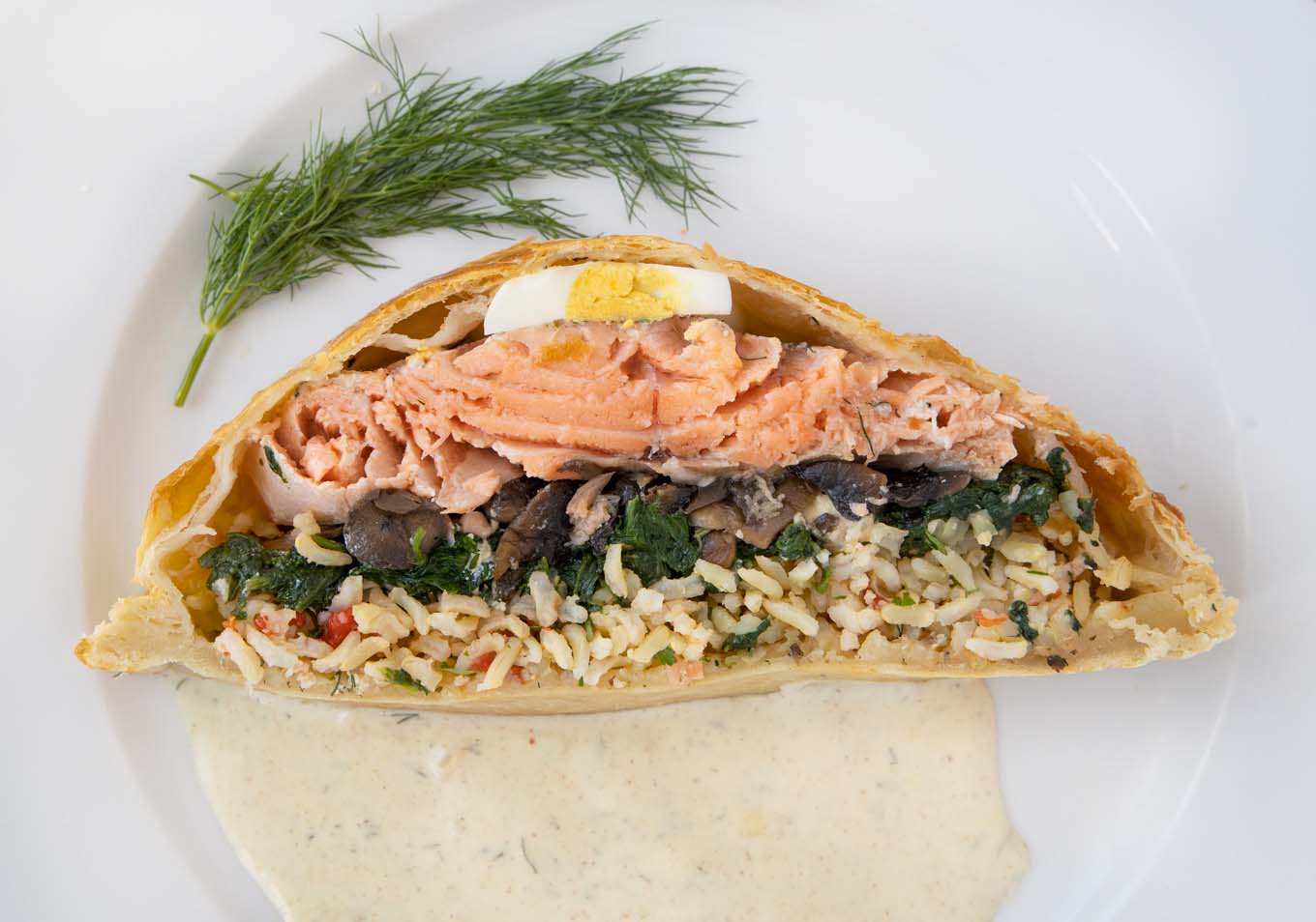 overhead view of a slice of Coulibiac of Salmon on a white plate with a dijon dill sauce and sprig of dill