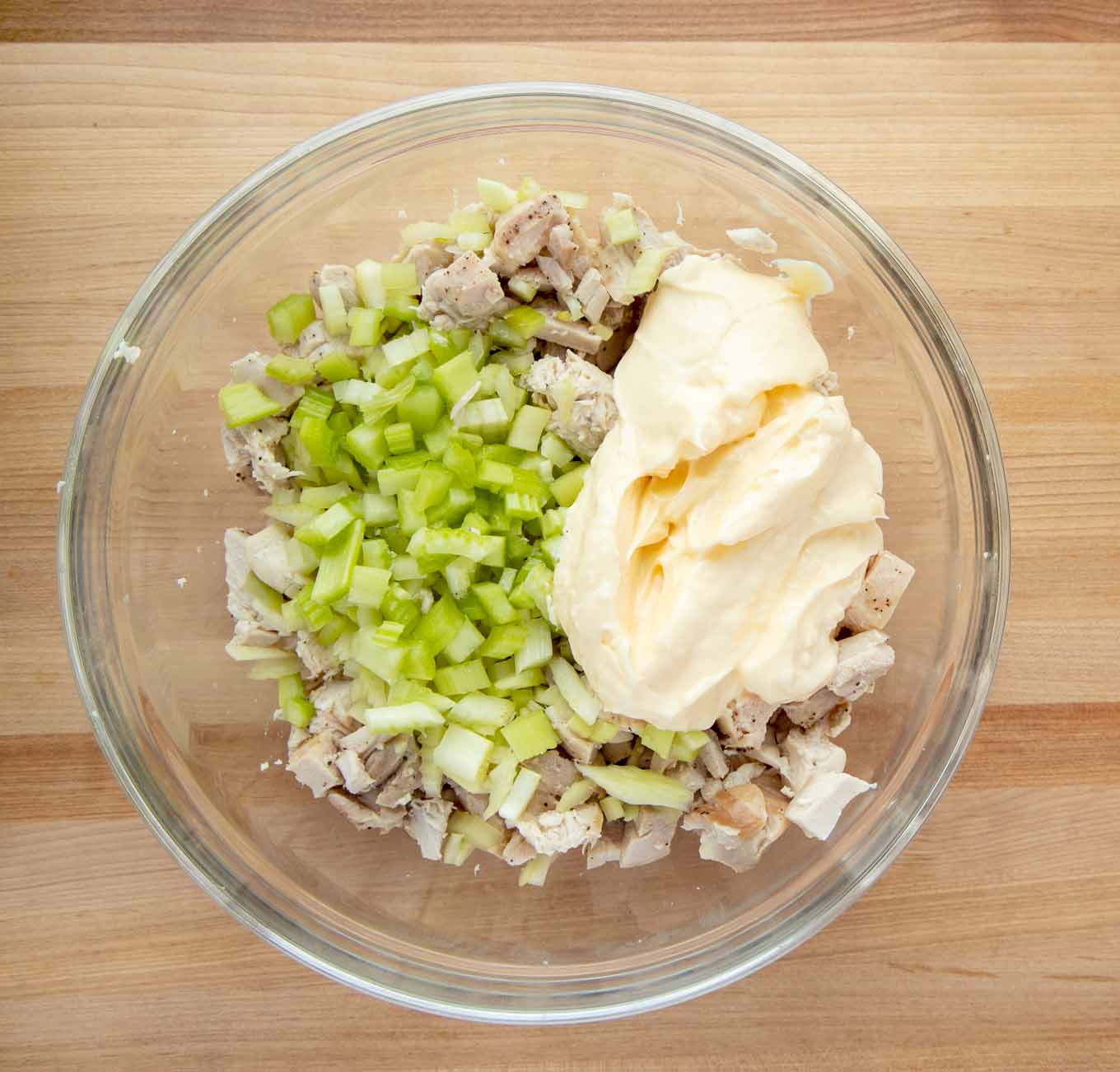 overhead view of chopped chicken, diced celery and mayonaise in a glass bowl on a wooden cutting board