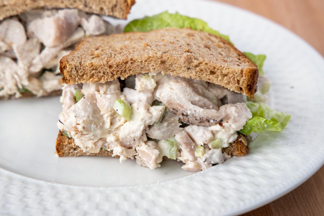 half of a chicken salad sandwich with lettuce on grain bread on a white plate with the other half behind it