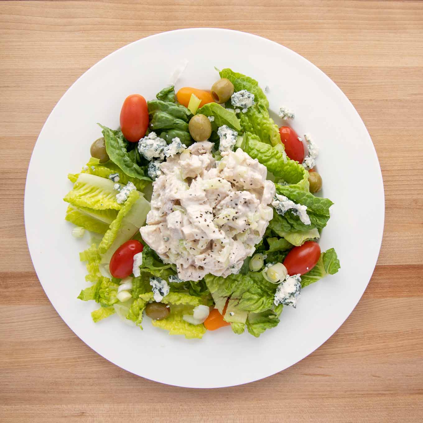 overhead view of salad on a white plate consisting of chopped romaine, grape tomatoes, green olives, gorgonzola cheese crumbles and big scoop of chicken salad in the middle