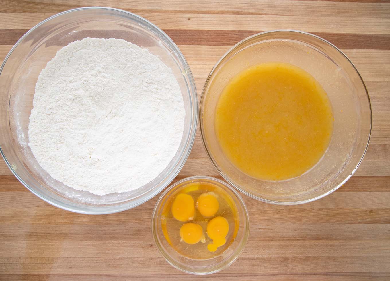 overhead view of a large bowl with flour mixture, a smaller bowl with oil, sugar mixture, a small bowl with eggs on a wooden cutting board
