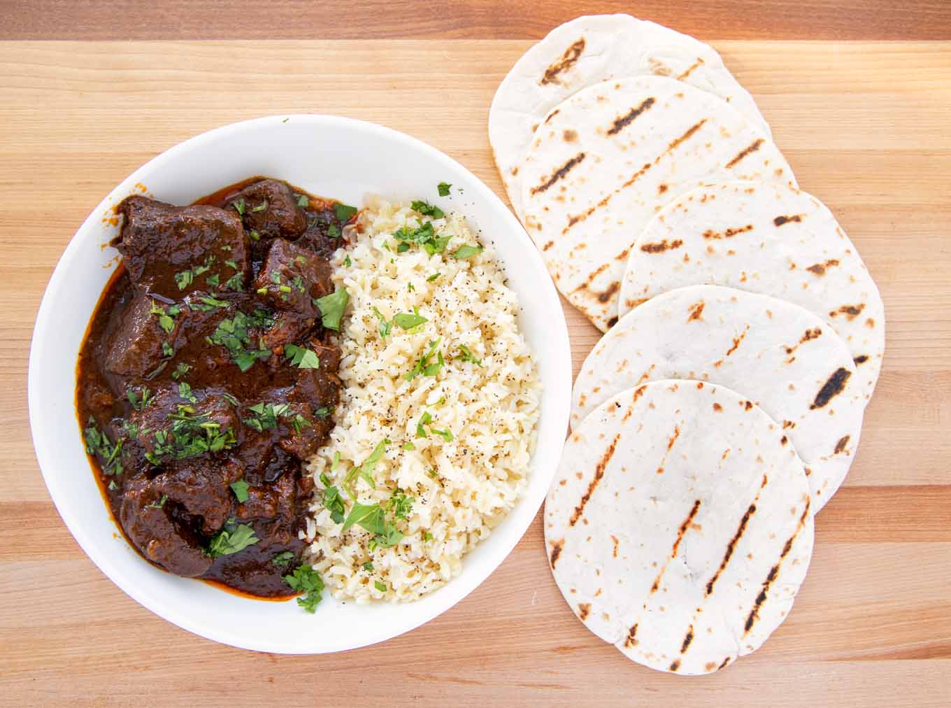 overhead view of a white bowl with beef chili colorado and rice with five grilled small flour tortillas on the side