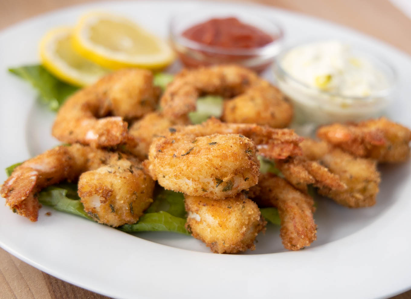 close up of golden brown fried shrimp on a white plate with tarter sauce, cocktail sauce and lemon slices