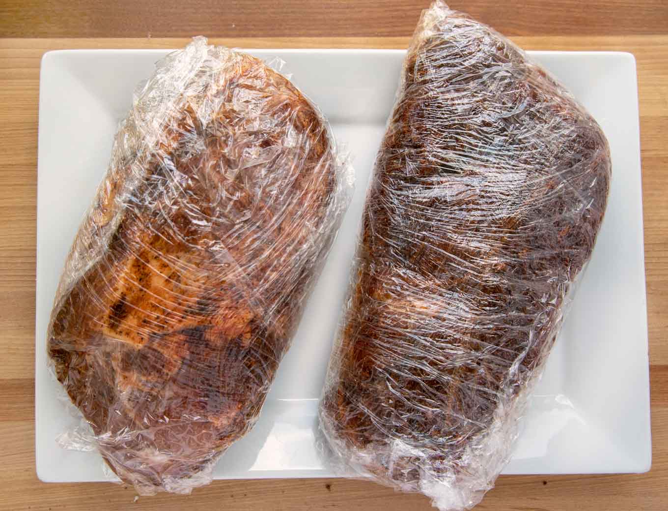 2 pork roasts coated with a dry rub and double wrapped in plastic wrap sitting on a white platter