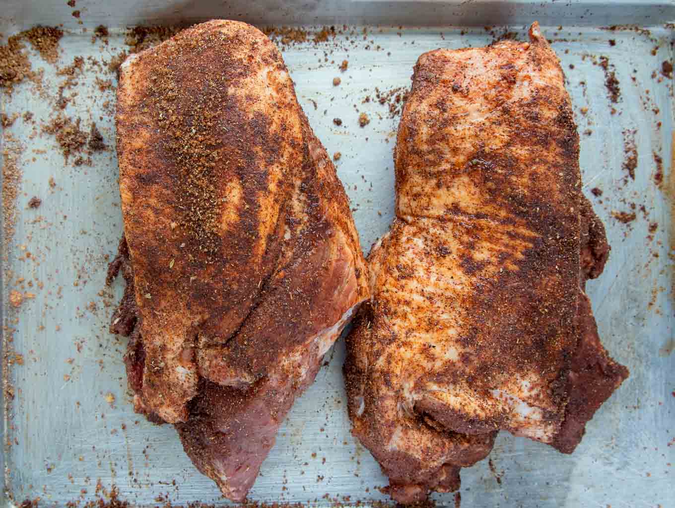 pork butt split in half, coated with a dry rub sitting on a sheet pan.