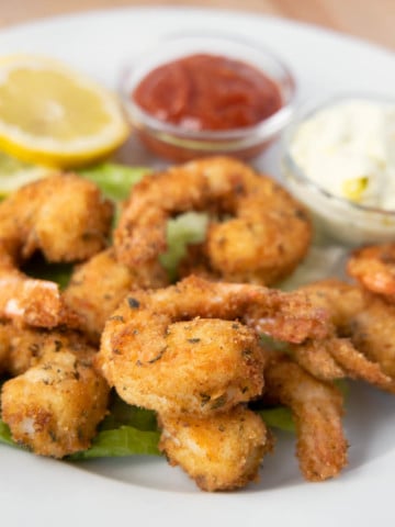 golden brown fried shrimp on a white plate with tarter sauce, cocktail sauce and lemon slices