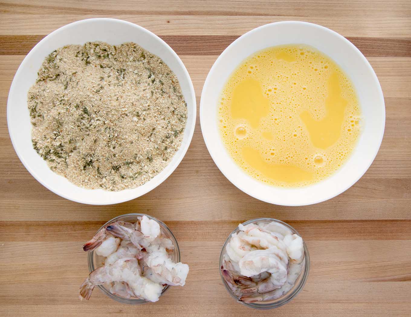 overhead view of raw shrimp and two white bowls with eggwash in one and seasoned bread crumbs in another