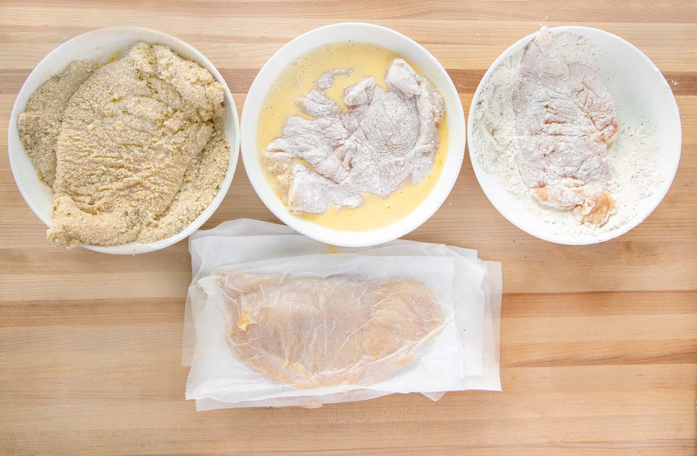 Chicken breasts with breading station with a piece of chicken in each stage of the breading station.