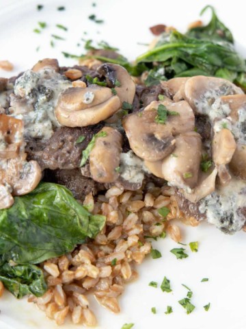 pan seared steak on a bed of farro topped with spinach mushrooms and blue cheese served on a white plate