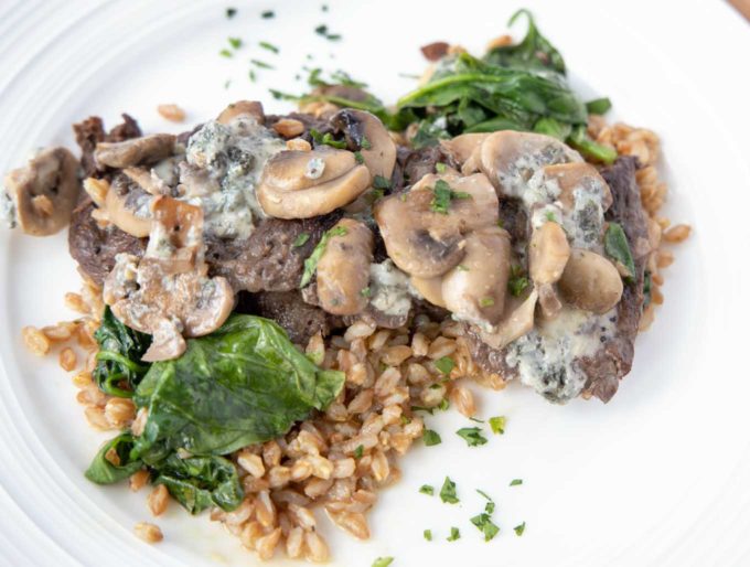 pan seared steak on a bed of farro topped with spinach mushrooms and blue cheese served on a white plate